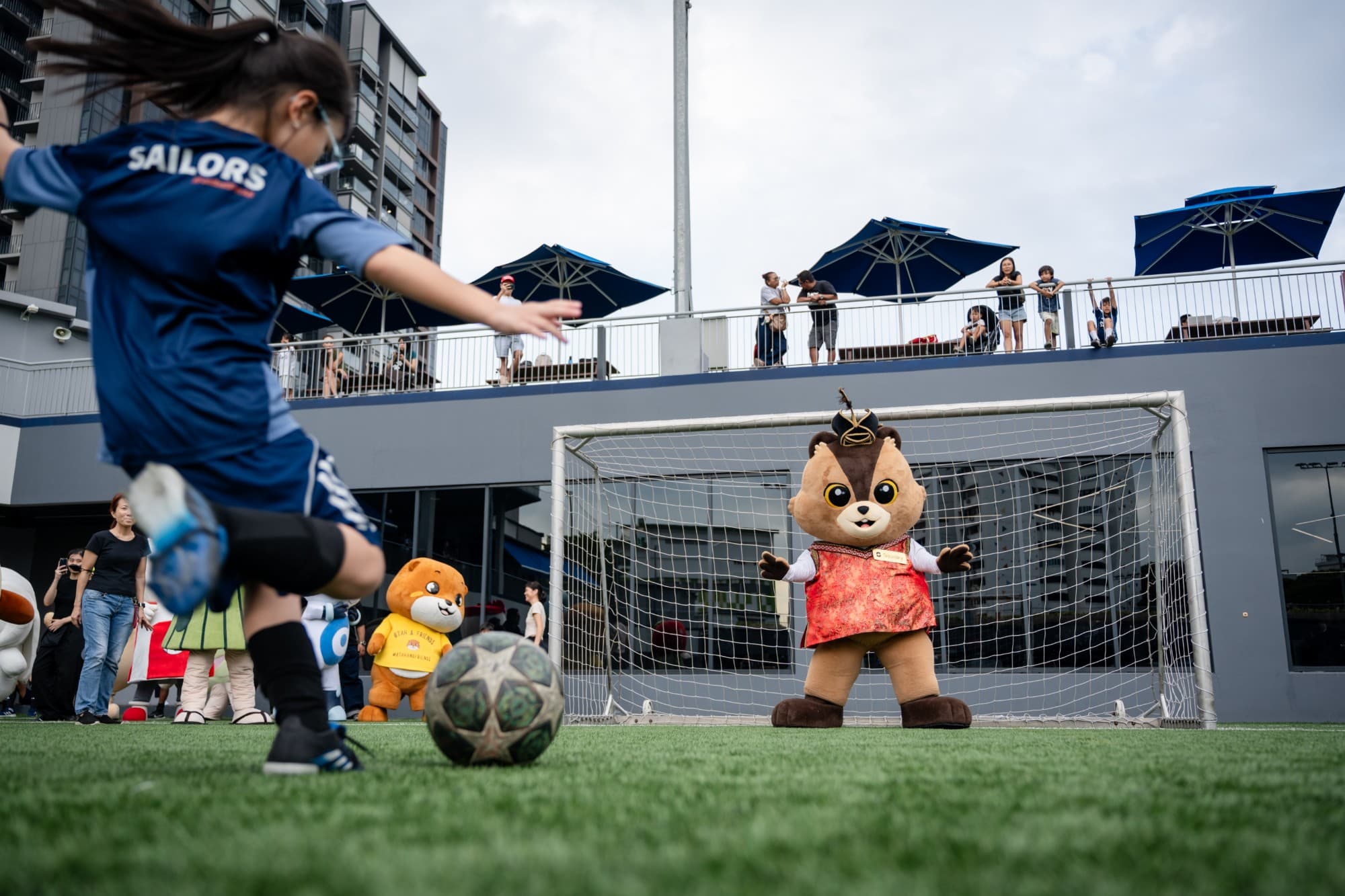 A girl takes a penalty kick against a bear mascot in goal at a football event