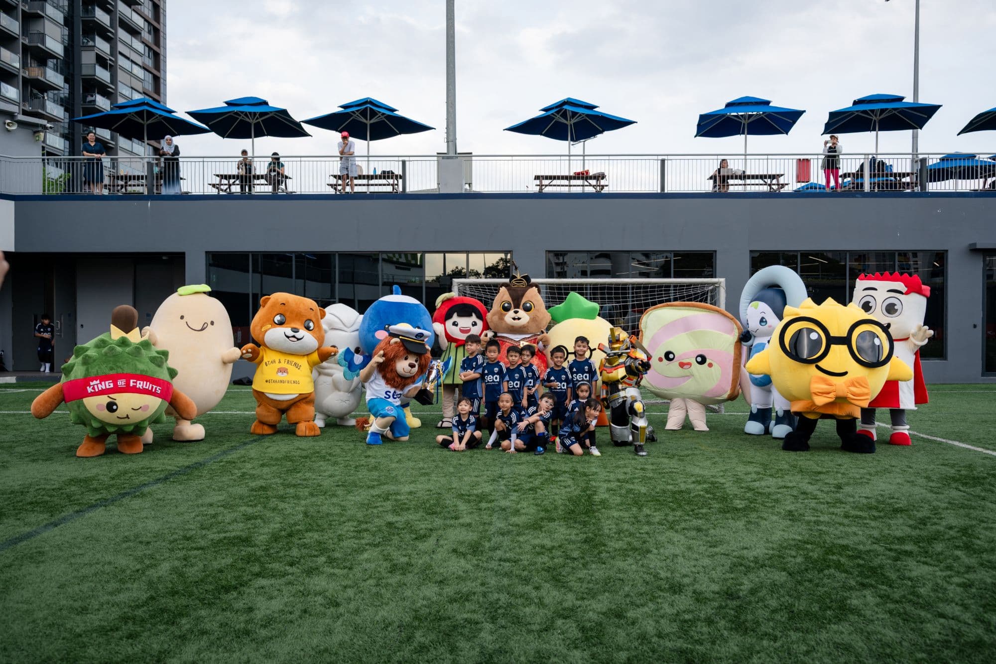 A line of mascots, including Otah and Friends, with a junior football team on the pitch